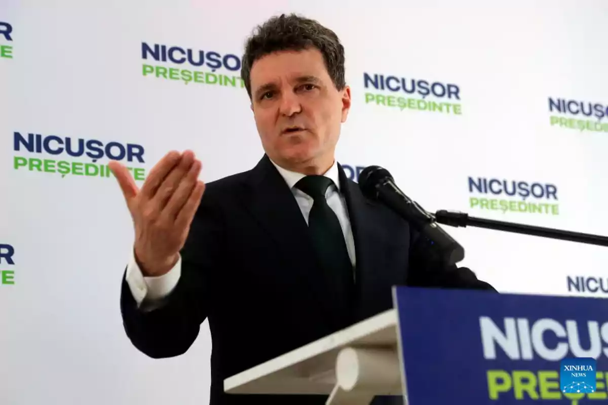 Un hombre en traje oscuro habla en un podio con un micrófono, con un fondo que tiene el texto Un hombre en traje oscuro habla en un podio con un micrófono, con un fondo que tiene el texto