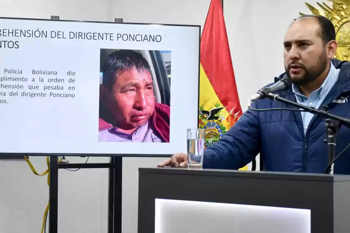 Un hombre hablando en una conferencia de prensa con una pantalla que muestra información sobre la aprehensión de un dirigente, junto a una bandera de Bolivia.