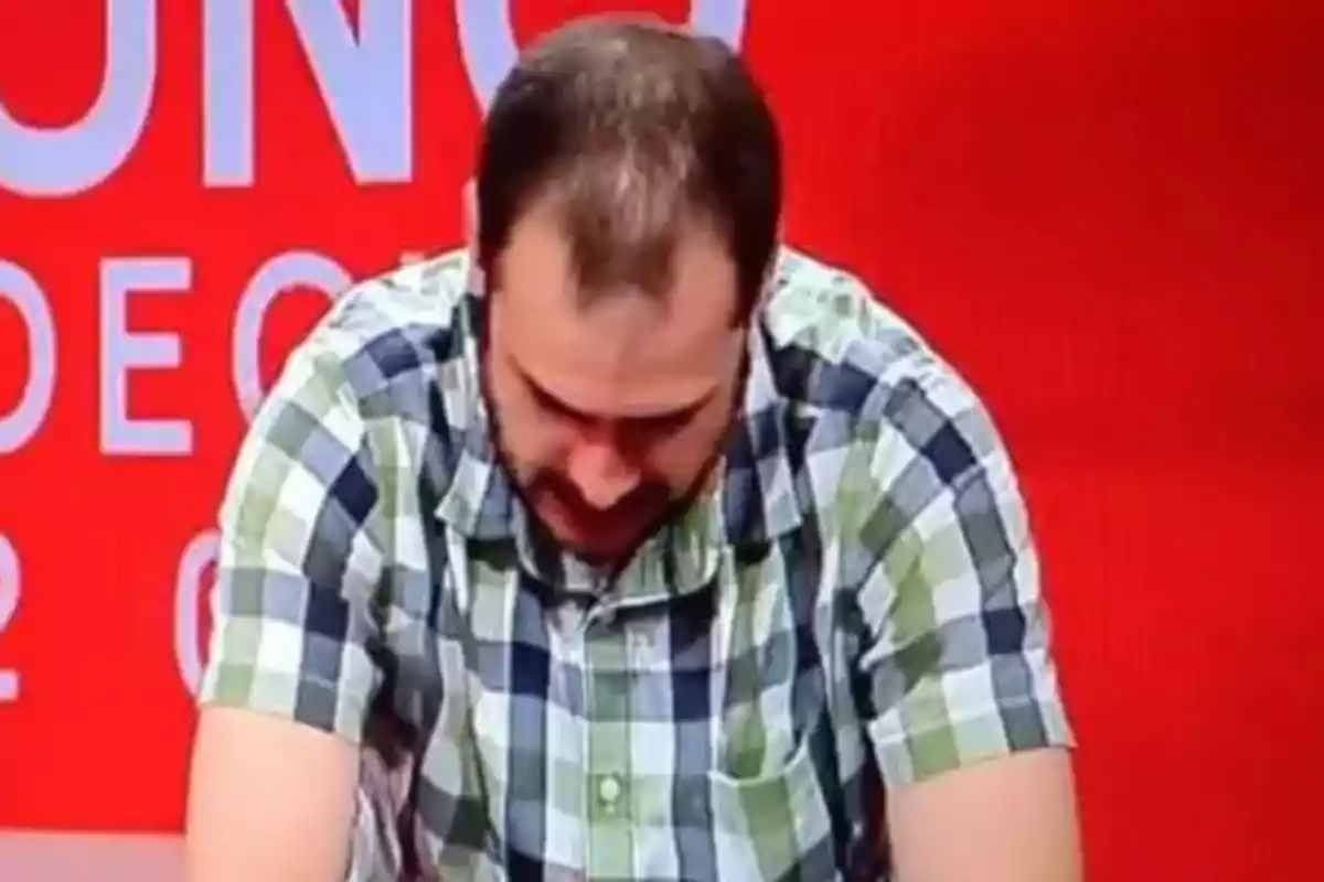 A man in a checkered jersey tilts his head down against a red background with white letters.