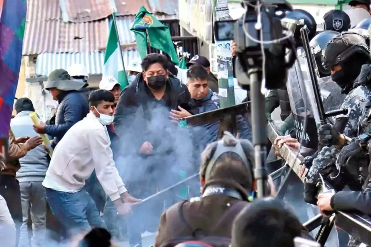 Personas manifestándose frente a un grupo de policías antidisturbios mientras hay humo en el ambiente y varias banderas ondean al fondo Personas manifestándose frente a un grupo de policías antidisturbios mientras hay humo en el ambiente y varias banderas ondean al fondo