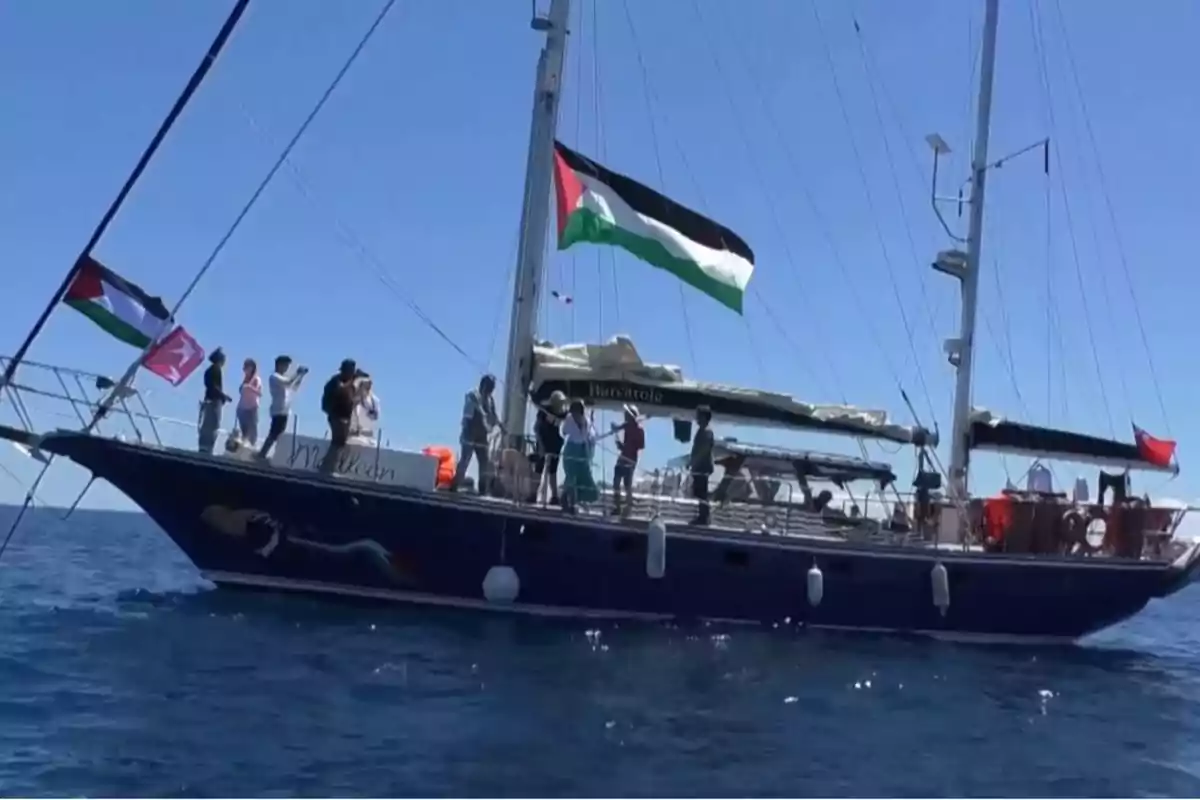 A blue sailboat sails on the sea with several people on board and Palestinian flags waving under a clear sky. A blue sailboat sails on the sea with several people on board and Palestinian flags waving under a clear sky.