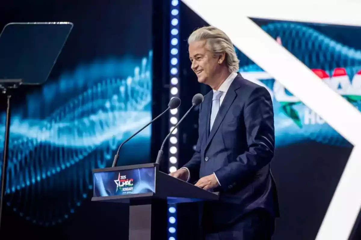 Hombre de cabello canoso y traje azul hablando en un podio durante un evento con fondo azul y luces brillantes Hombre de cabello canoso y traje azul hablando en un podio durante un evento con fondo azul y luces brillantes
