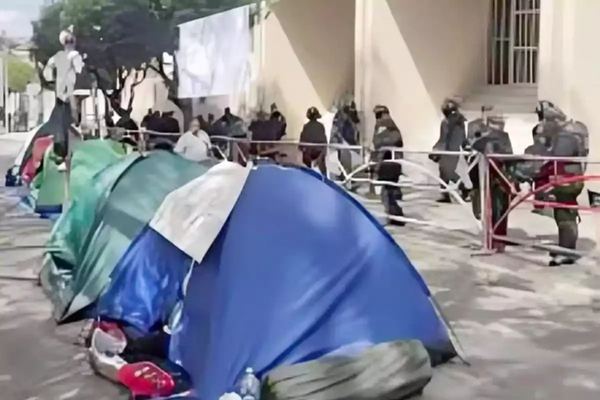 Varias casas de campaña alineadas en una acera con personas y policías al fondo junto a una barrera metálica Varias casas de campaña alineadas en una acera con personas y policías al fondo junto a una barrera metálica