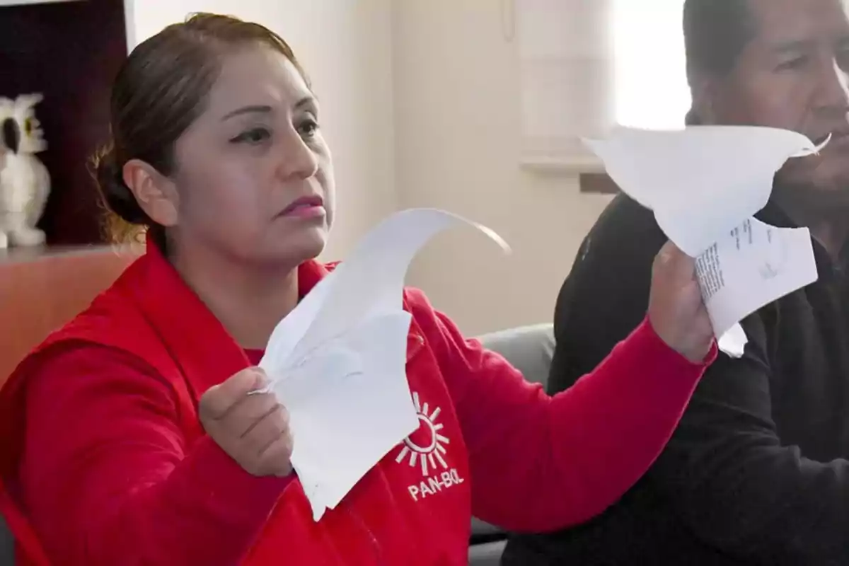 Mujer con chaqueta roja rasgando hojas de papel mientras está sentada junto a otra persona