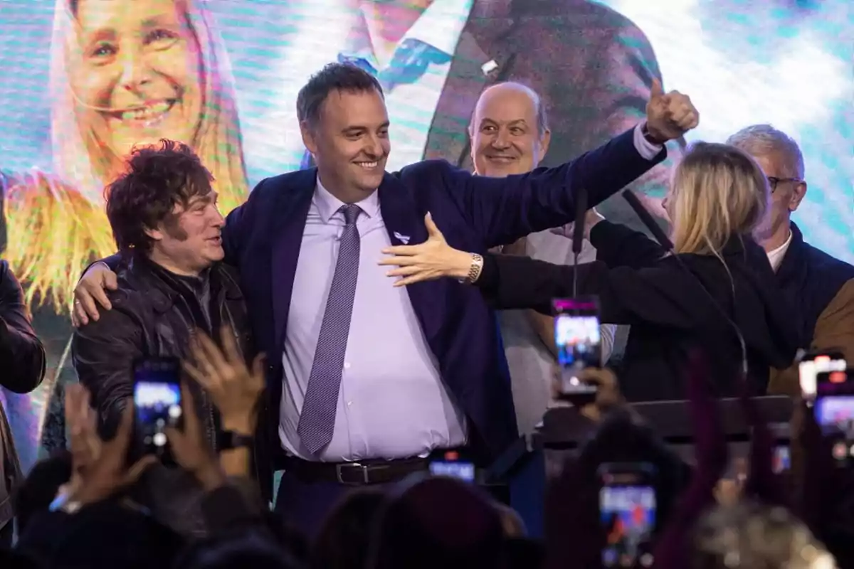 A group of people smiles and celebrates on a stage while being photographed by the audience. A group of people smiles and celebrates on a stage while being photographed by the audience.