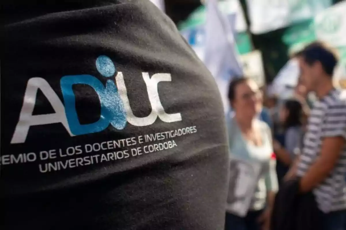 Persona con camiseta negra de ADIUC en primer plano mientras al fondo se observa un grupo de personas en una manifestación o reunión al aire libre