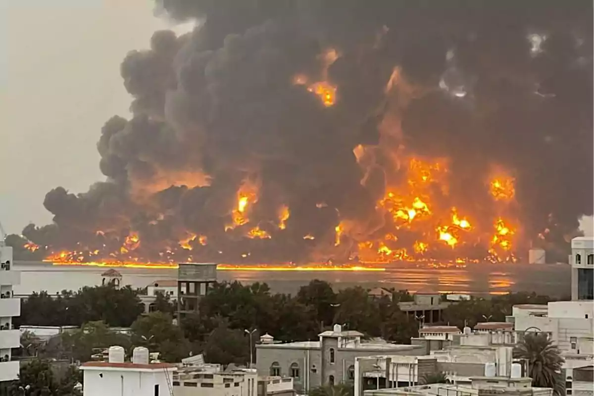 Un gran incendio con llamas intensas y humo denso se eleva sobre una zona urbana cercana al agua.