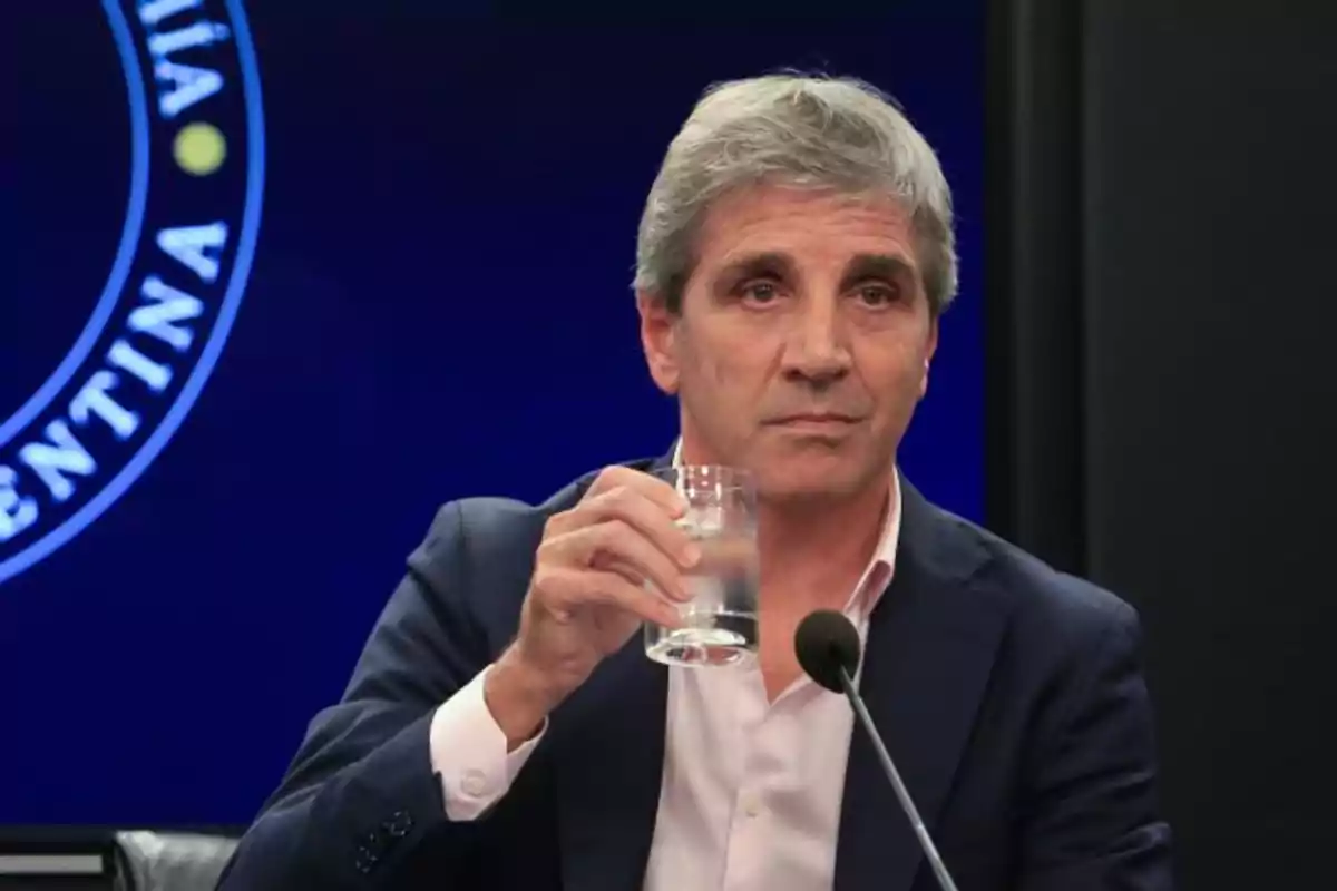 Un hombre de cabello canoso sostiene un vaso de agua mientras está sentado frente a un micrófono con un fondo azul que tiene un texto circular.