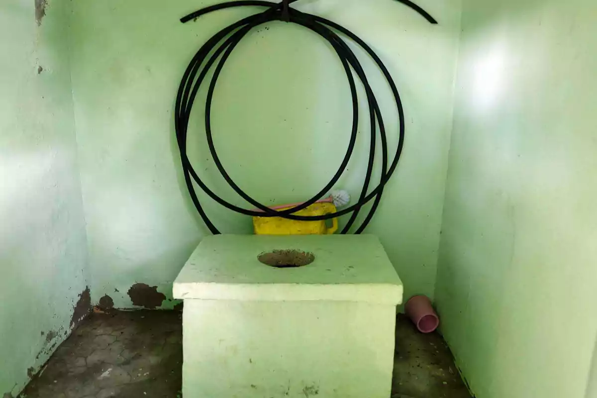 Baño rústico de letrina con paredes verdes, un tubo enrollado en la pared y un balde amarillo con escobilla detrás Baño rústico de letrina con paredes verdes, un tubo enrollado en la pared y un balde amarillo con escobilla detrás