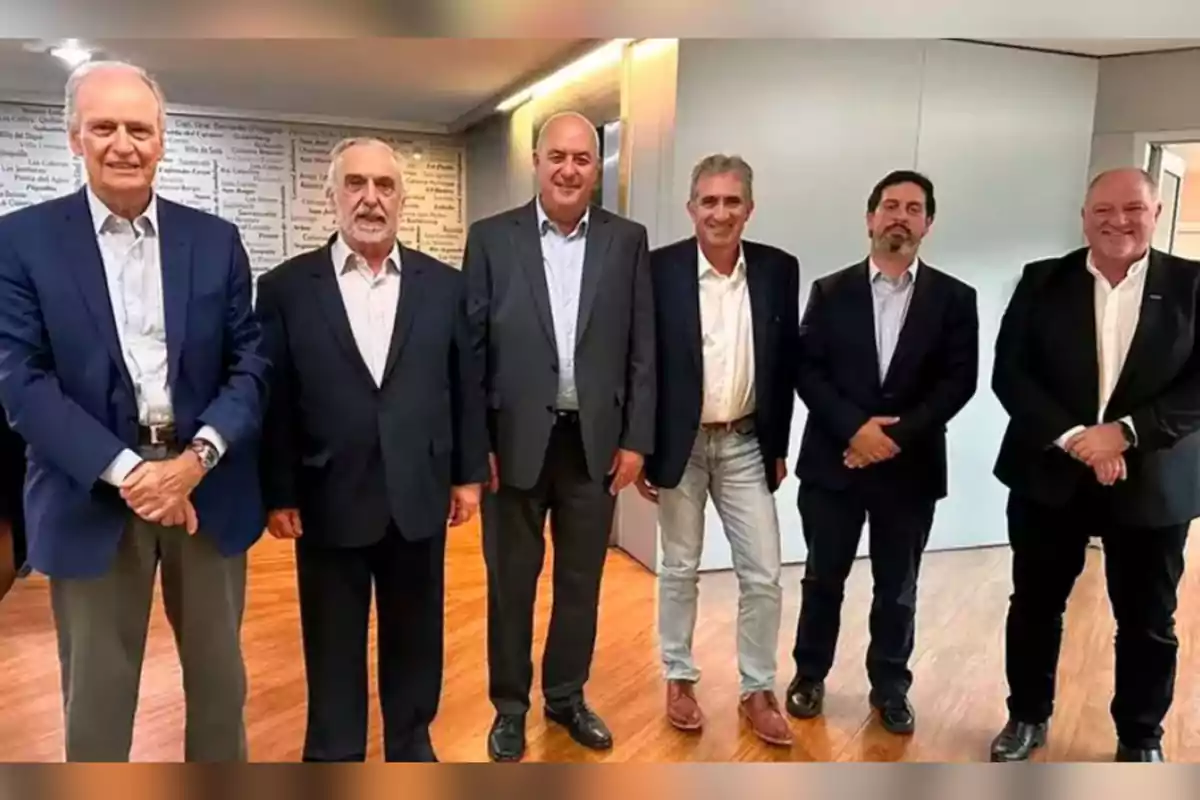 Seis hombres vestidos con traje posan juntos en una oficina con piso de madera y paredes claras