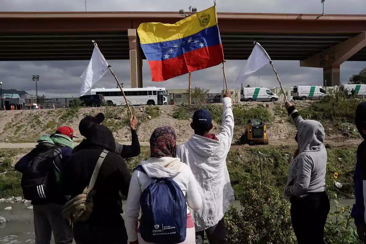 A group of people holds white flags and a Venezuelan flag while observing a border area with official vehicles and a bridge in the background. A group of people holds white flags and a Venezuelan flag while observing a border area with official vehicles and a bridge in the background.