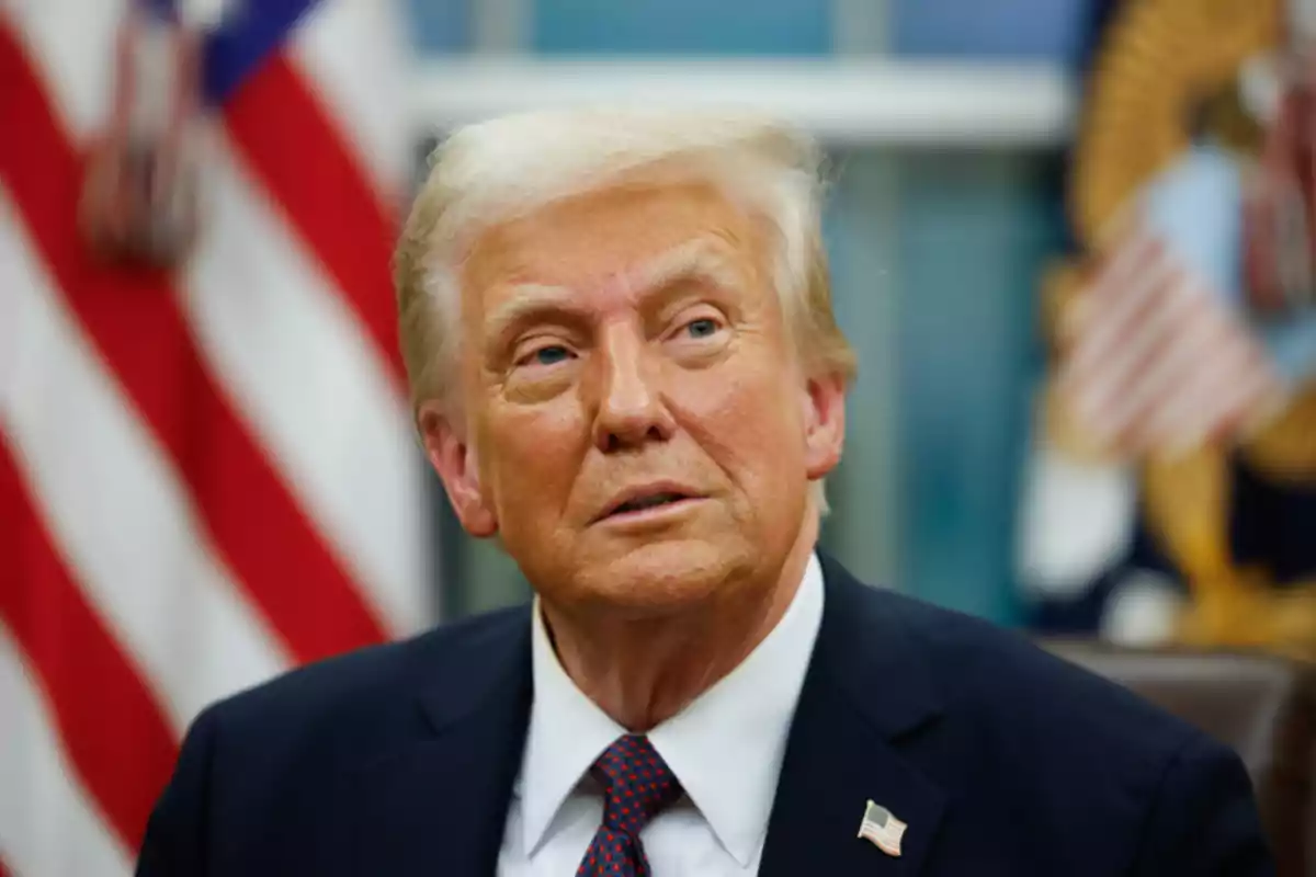 Un hombre de cabello rubio y traje oscuro con una corbata roja, sentado frente a banderas estadounidenses. Un hombre de cabello rubio y traje oscuro con una corbata roja, sentado frente a banderas estadounidenses.