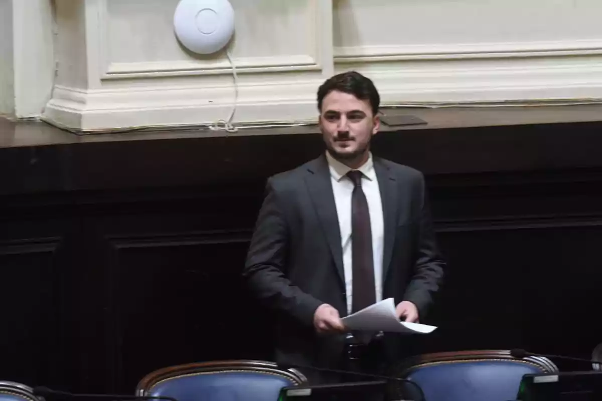 Un hombre de traje y corbata sostiene papeles mientras está de pie en una sala con sillas y una pared decorativa.