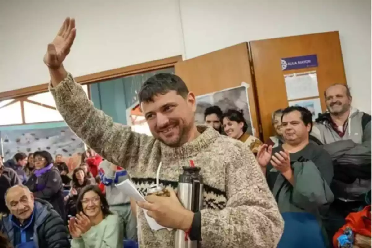 Un hombre sonriente levanta la mano mientras sostiene un termo y una hoja de papel rodeado de personas que aplauden en una sala.