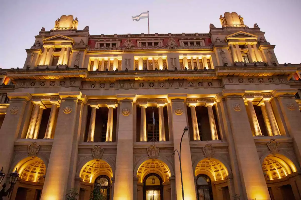 Fachada iluminada de un edificio histórico con columnas y una bandera argentina en la parte superior Fachada iluminada de un edificio histórico con columnas y una bandera argentina en la parte superior