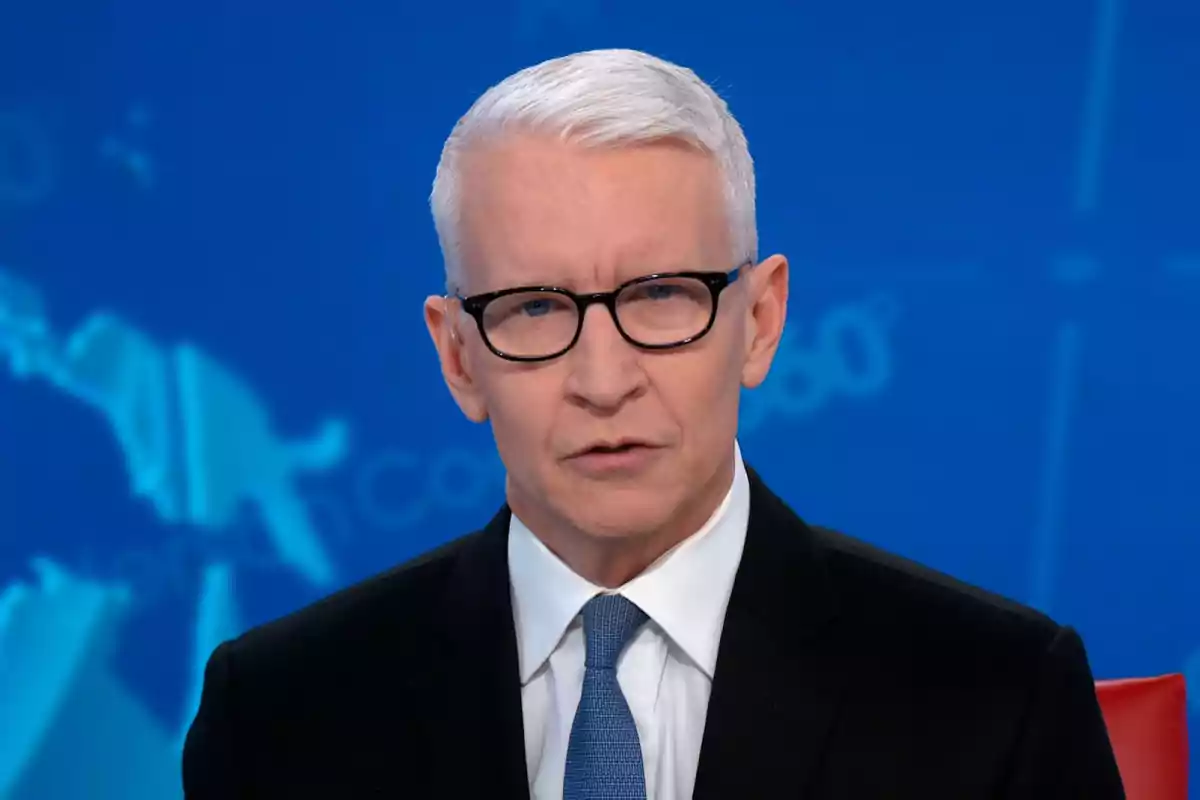 A gray-haired man with glasses is speaking in front of a camera with a blue background. A gray-haired man with glasses is speaking in front of a camera with a blue background.