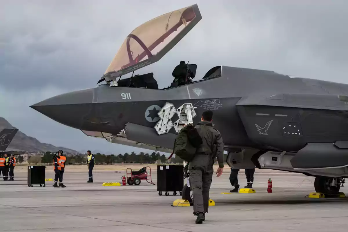 Un piloto camina hacia un avión de combate en una pista de aterrizaje mientras varias personas con chalecos naranjas están en el fondo. Un piloto camina hacia un avión de combate en una pista de aterrizaje mientras varias personas con chalecos naranjas están en el fondo.