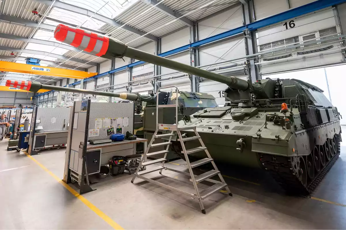 Un tanque militar en un taller de mantenimiento con una escalera de acceso y equipo de trabajo alrededor.