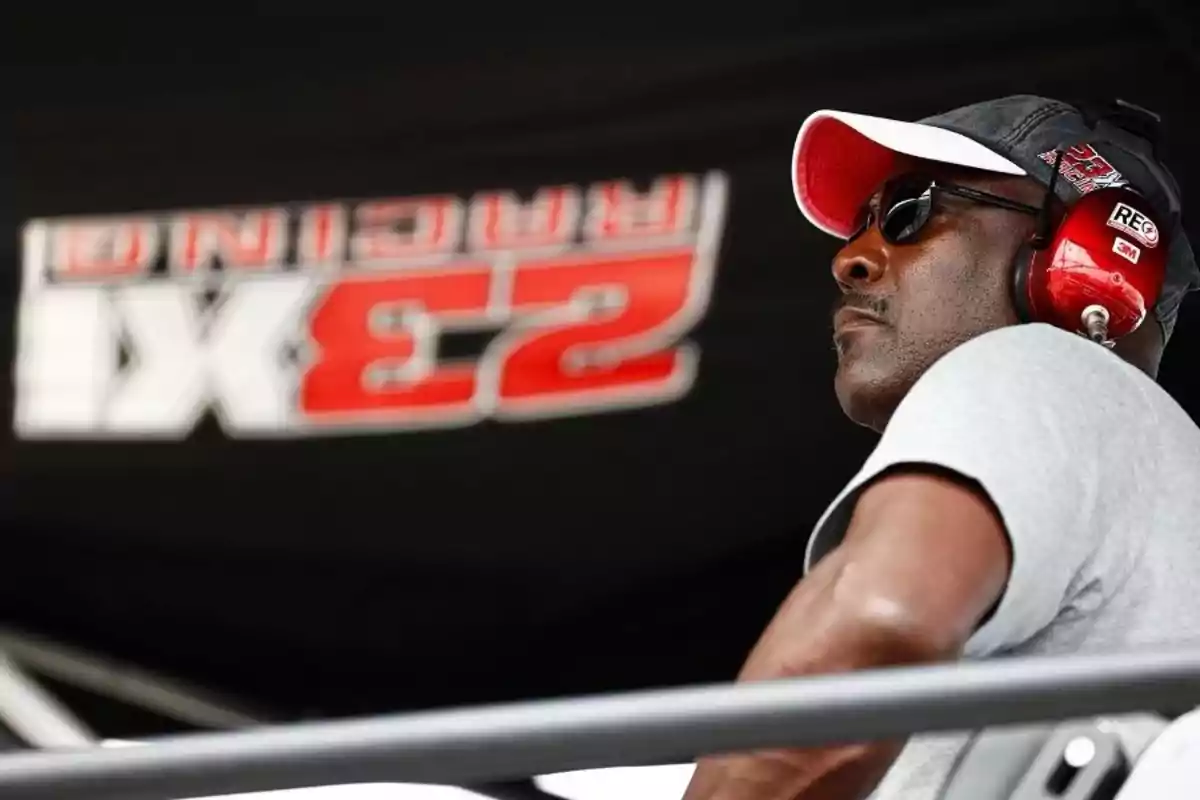 Un hombre con gafas de sol y auriculares rojos observa atentamente en un entorno relacionado con carreras, con un fondo que muestra un logotipo de equipo de automovilismo. Un hombre con gafas de sol y auriculares rojos observa atentamente en un entorno relacionado con carreras, con un fondo que muestra un logotipo de equipo de automovilismo.
