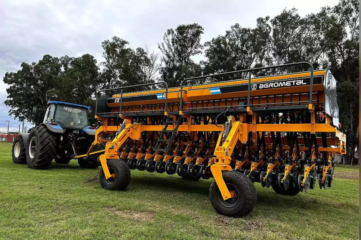 Tractor azul enganchado a una sembradora agrícola amarilla de gran tamaño sobre césped con árboles al fondo Tractor azul enganchado a una sembradora agrícola amarilla de gran tamaño sobre césped con árboles al fondo