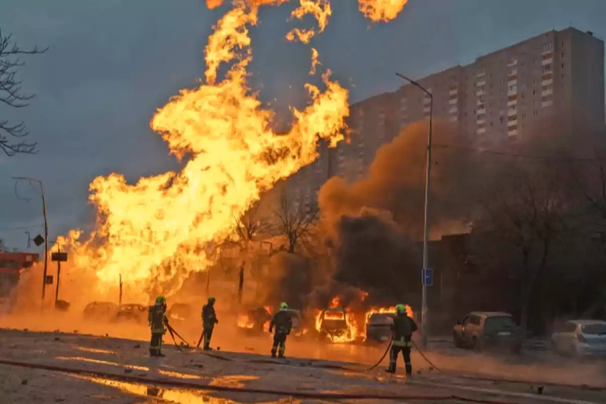 Bomberos combaten un gran incendio en una calle con varios autos en llamas y una enorme columna de fuego y humo frente a un edificio residencial Bomberos combaten un gran incendio en una calle con varios autos en llamas y una enorme columna de fuego y humo frente a un edificio residencial