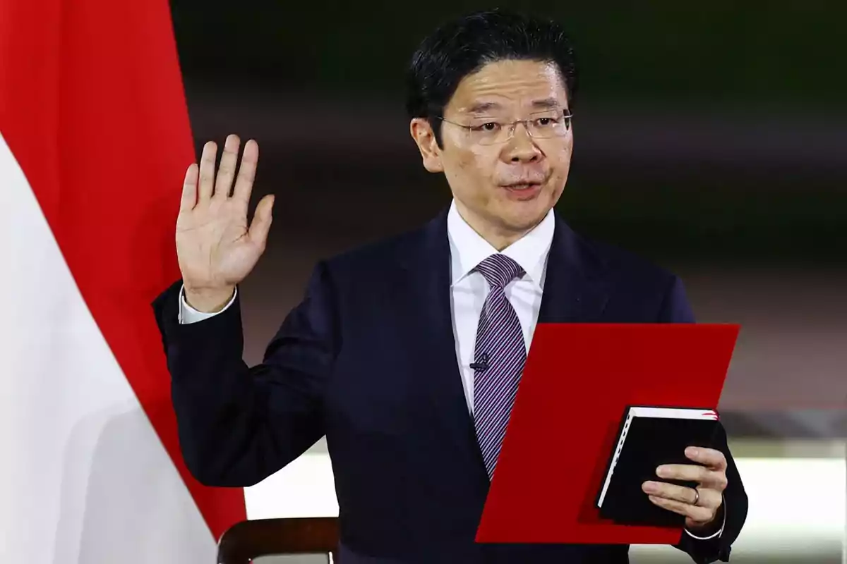 A man in a dark suit and striped tie holds a red document and a notebook while raising his hand in front of a flag. A man in a dark suit and striped tie holds a red document and a notebook while raising his hand in front of a flag.