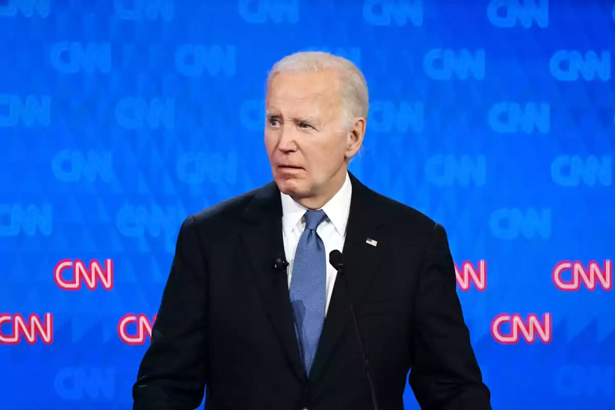 Gray-haired man in a dark suit and blue tie speaking in front of a blue background with CNN logos