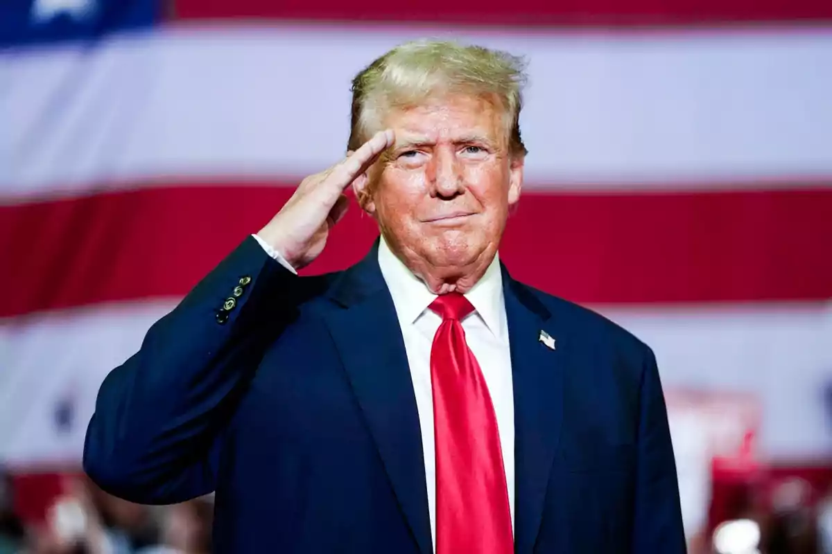 Un hombre de traje oscuro y corbata roja hace un saludo militar frente a una bandera de Estados Unidos.