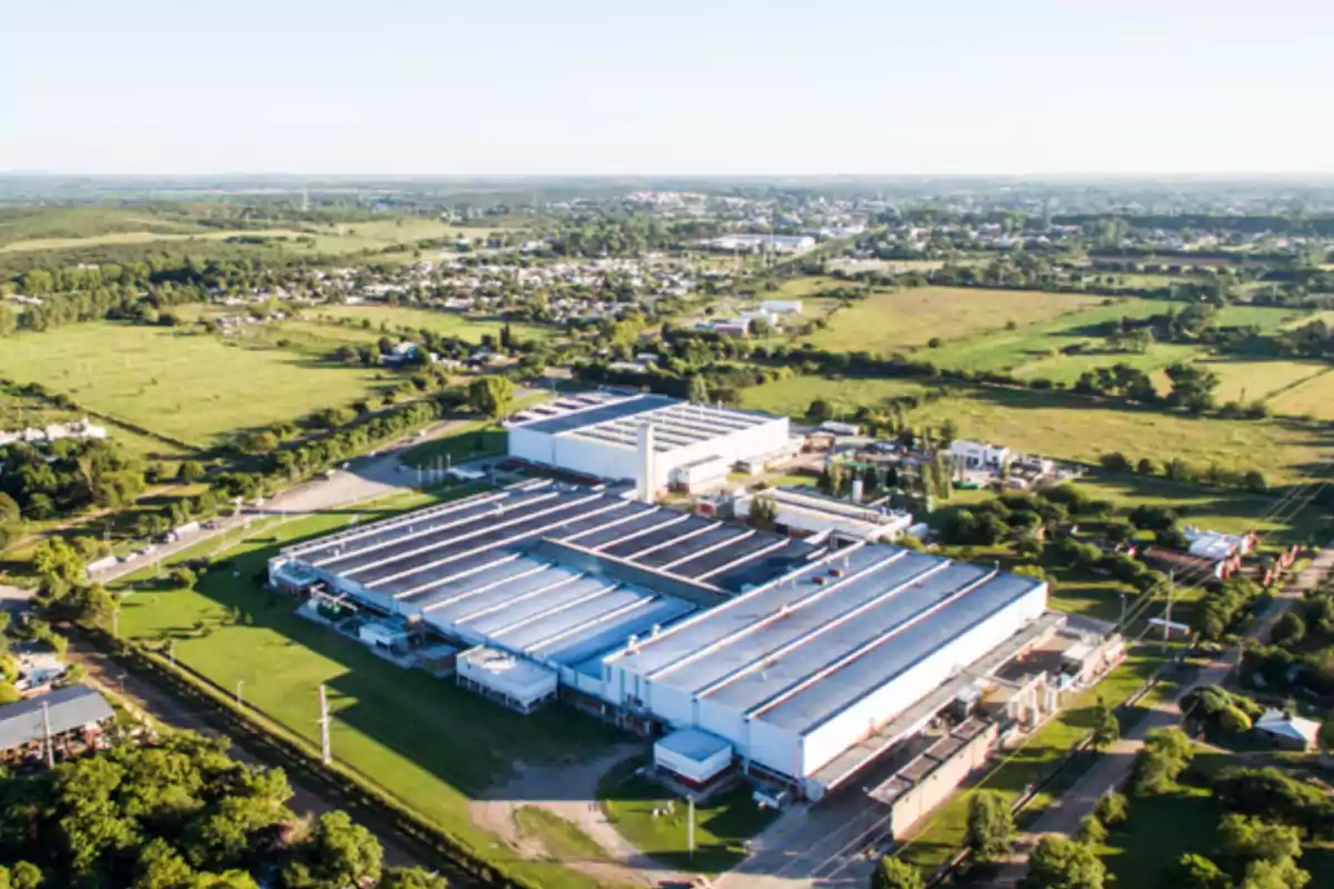 Vista aérea de una gran planta industrial rodeada de áreas verdes y campos con una ciudad al fondo Vista aérea de una gran planta industrial rodeada de áreas verdes y campos con una ciudad al fondo