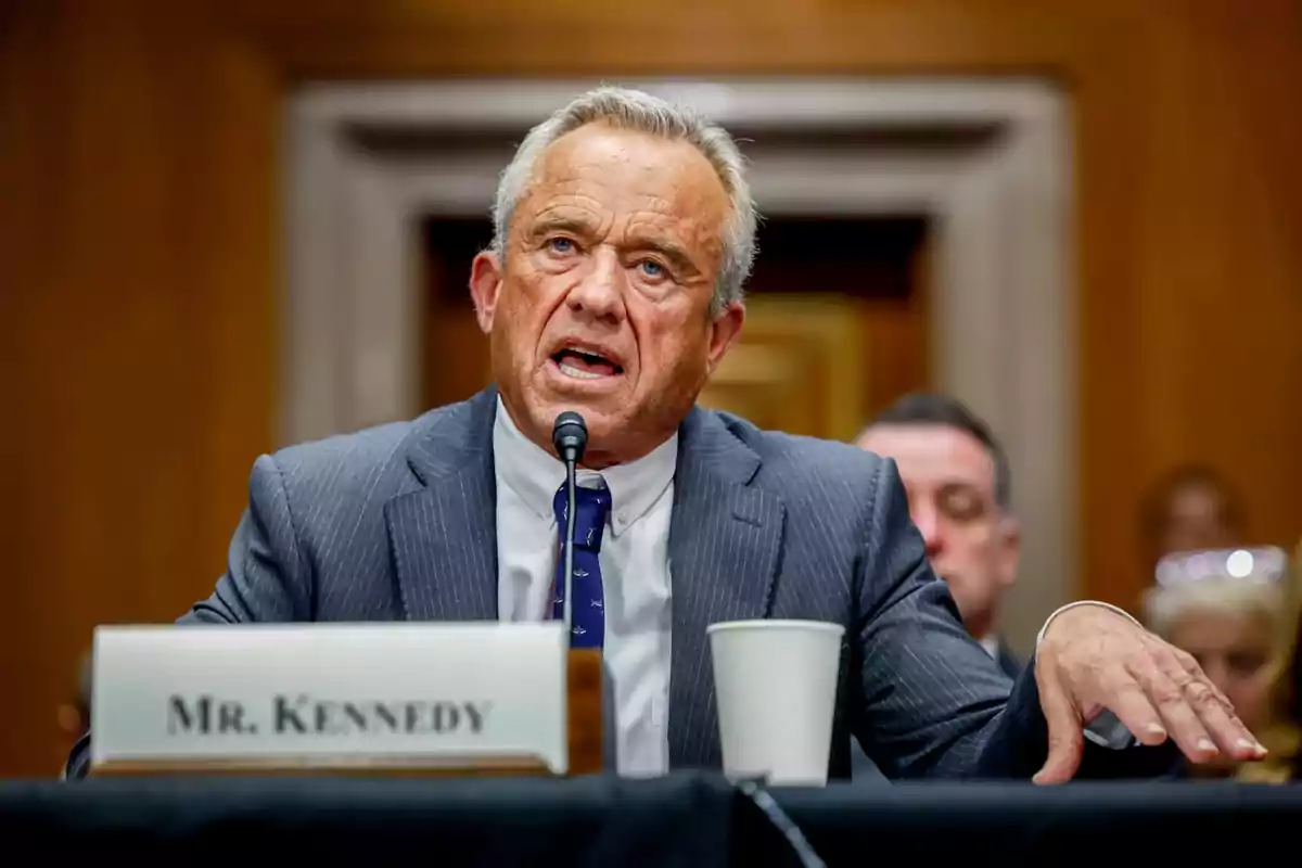 A man in a suit speaks into a microphone during a hearing in a formal setting.