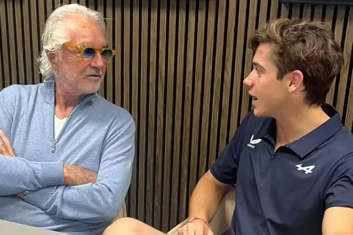 Dos hombres sentados conversan frente a una pared de madera, uno lleva gafas y suéter azul claro y el otro viste una camiseta azul marino con logotipo blanco. Dos hombres sentados conversan frente a una pared de madera, uno lleva gafas y suéter azul claro y el otro viste una camiseta azul marino con logotipo blanco.