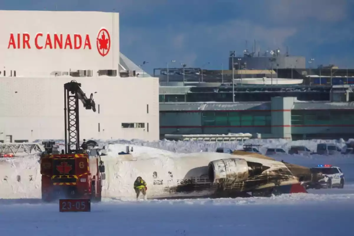 Un avión accidentado yace en la nieve cerca de un edificio de Air Canada, mientras un camión de bomberos y personal de emergencia están presentes en la escena.