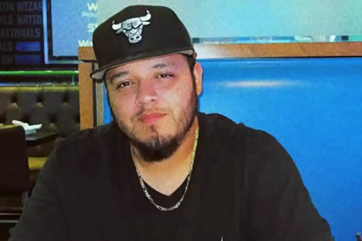 A man with a beard and a black cap with a bull logo is sitting in a restaurant with a blue background. A man with a beard and a black cap with a bull logo is sitting in a restaurant with a blue background.