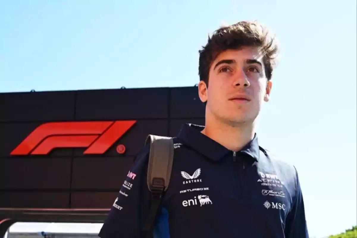 A young man in a racing team uniform stands in front of a Formula 1 poster under a clear sky.