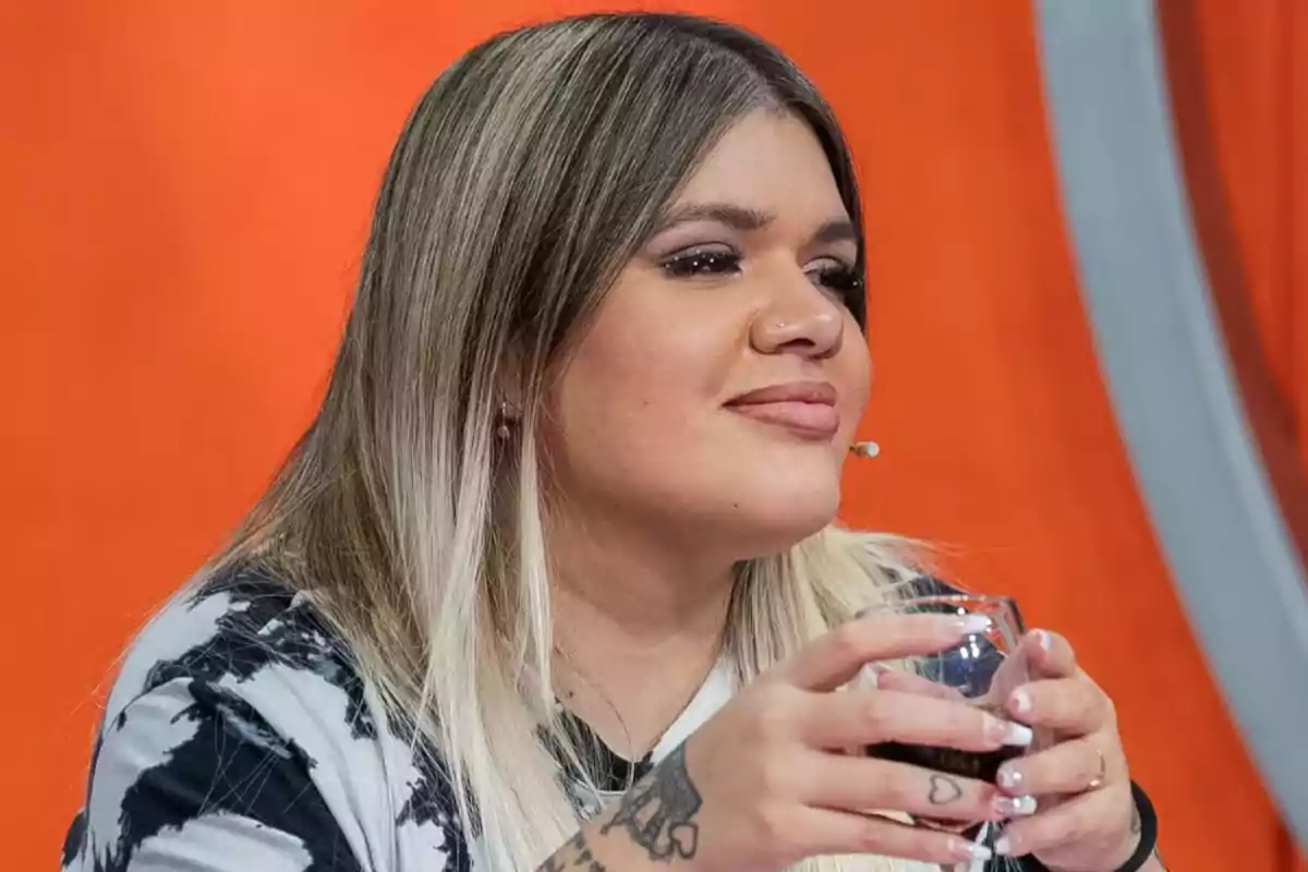 Una persona con cabello rubio y largo sostiene un vaso mientras está sentada frente a un fondo naranja.