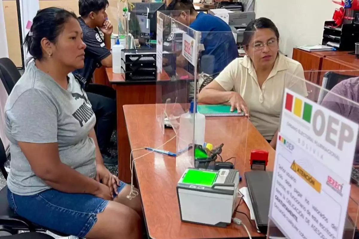 Una mujer está sentada en una oficina frente a un escritorio con un dispositivo de huellas digitales, mientras otra persona está del otro lado del escritorio, y hay un cartel del OEP en primer plano.