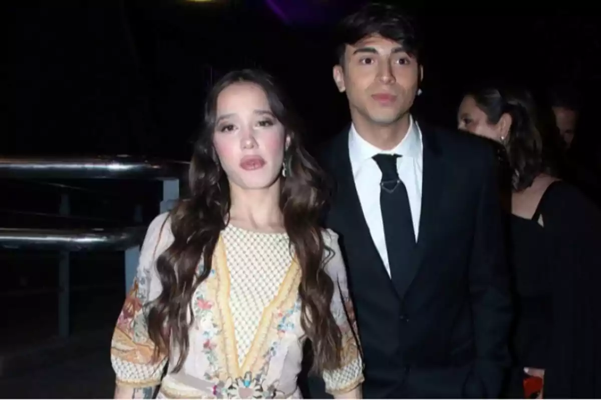 A young couple poses for the camera at a nighttime event; she is wearing a light-colored embroidered dress and he is dressed in a black suit with a tie.