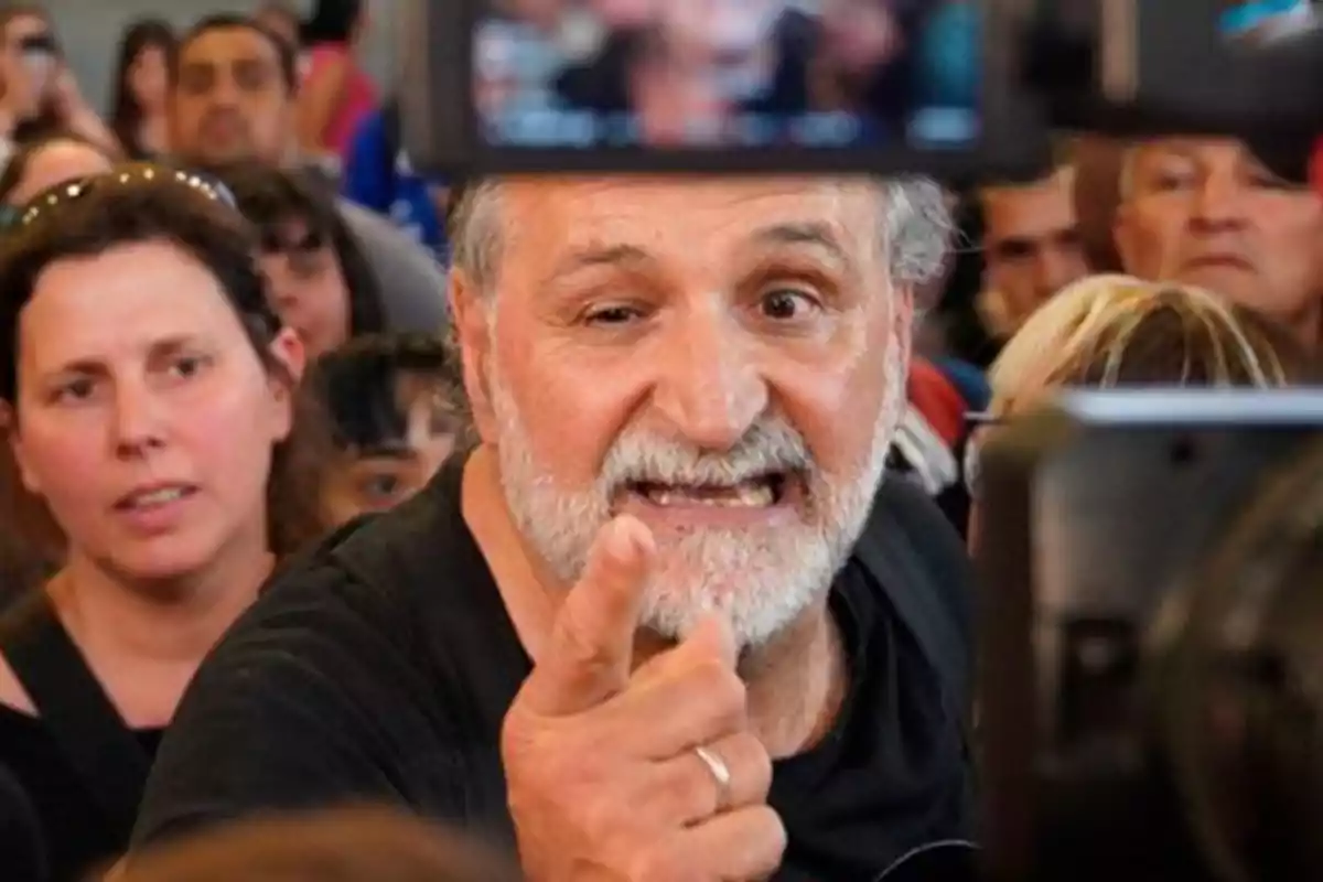 Un hombre de barba blanca gesticula y señala con el dedo en medio de una multitud.