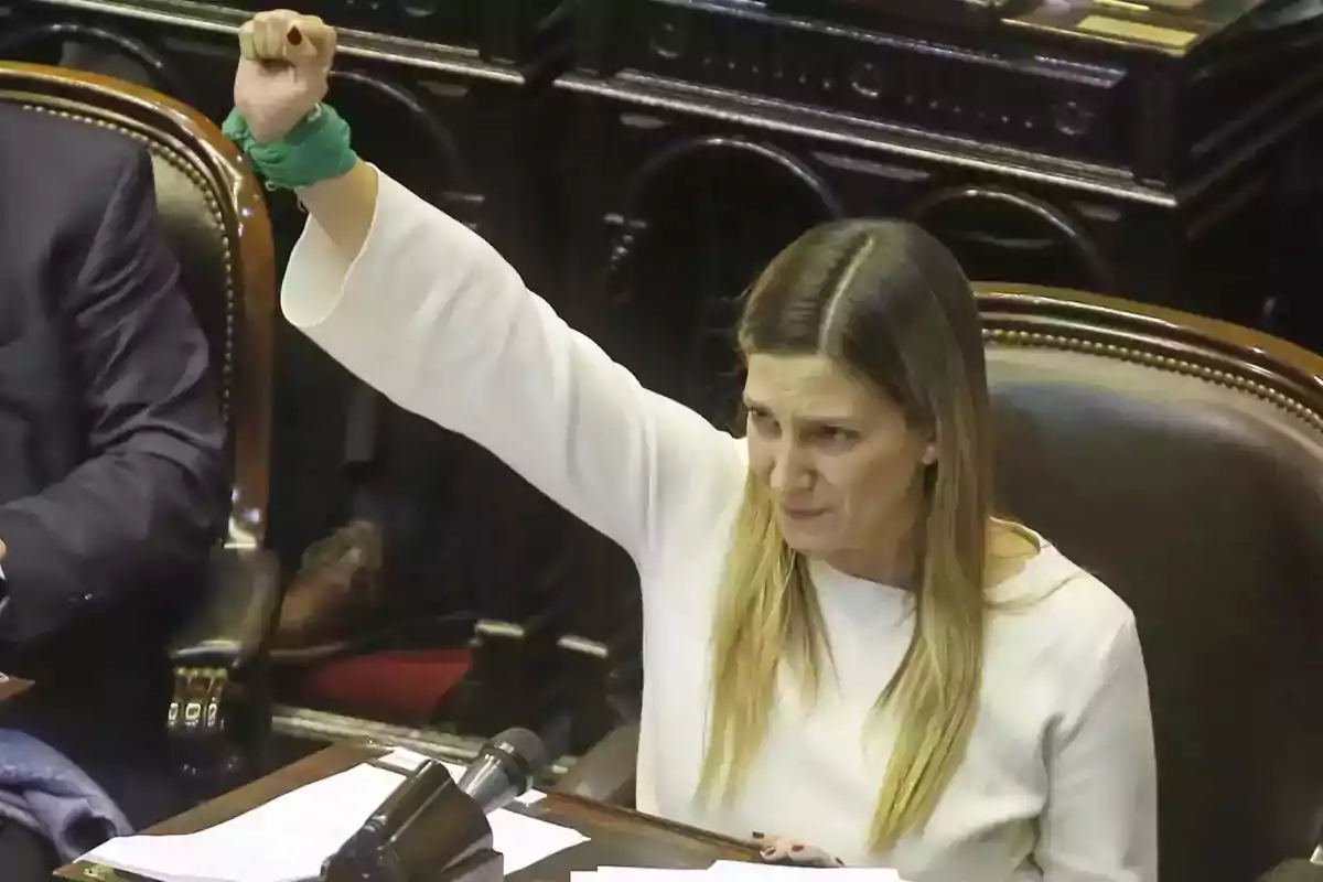 Mujer sentada en un recinto parlamentario levantando el puño derecho con un pañuelo verde en la muñeca Mujer sentada en un recinto parlamentario levantando el puño derecho con un pañuelo verde en la muñeca