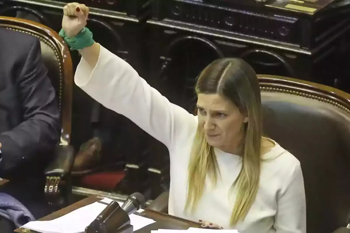 A person with their arm raised and a green handkerchief on their wrist, sitting in a formal setting with papers and a microphone in front of them. A person with their arm raised and a green handkerchief on their wrist, sitting in a formal setting with papers and a microphone in front of them.