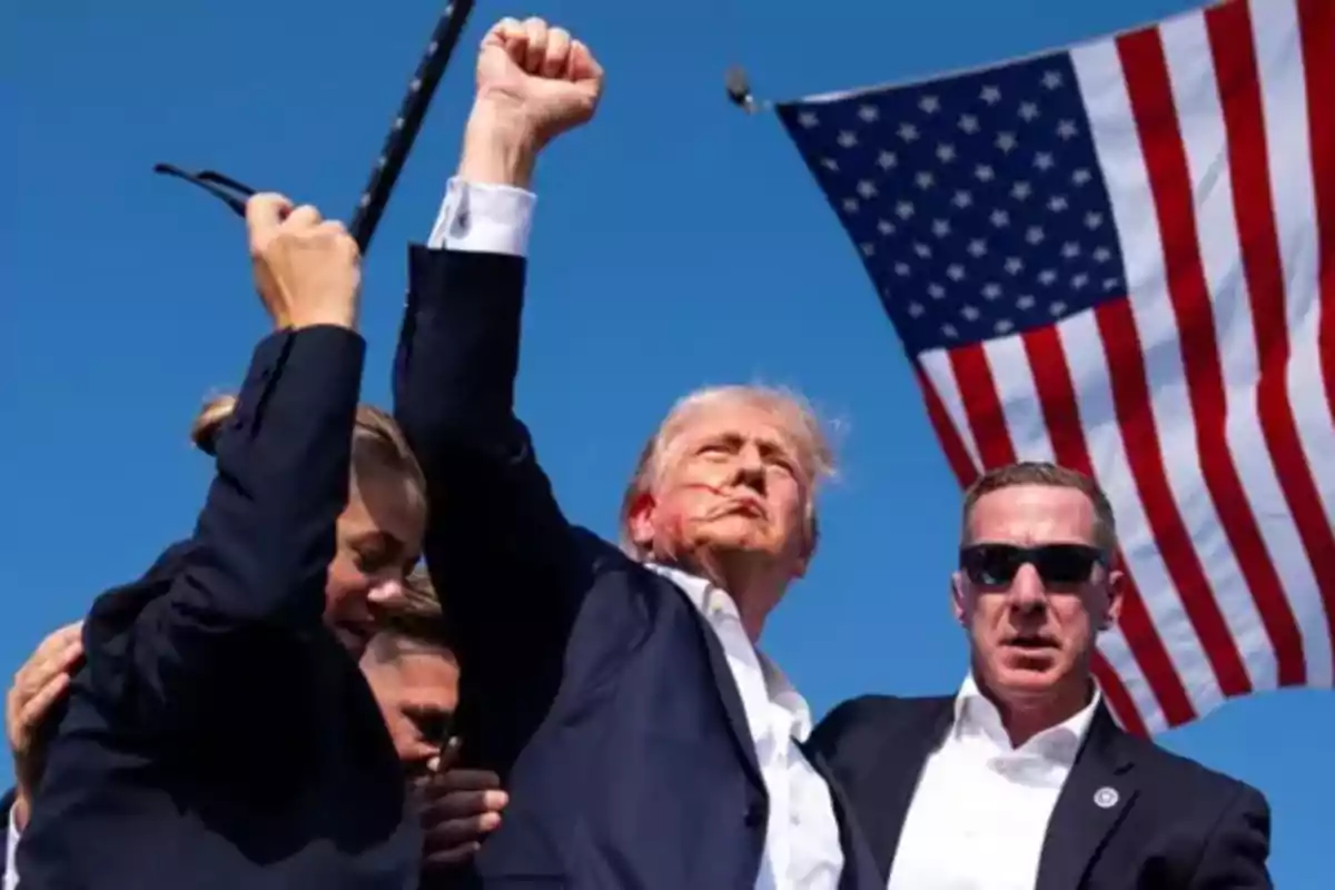 Un grupo de personas con trajes oscuros levanta el puño frente a una bandera de Estados Unidos ondeando en un cielo despejado.