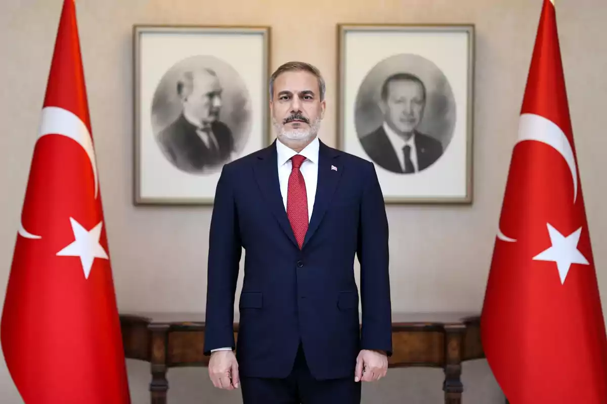 A man in a dark suit and red tie stands between two Turkish flags with black and white portraits on the wall behind him. A man in a dark suit and red tie stands between two Turkish flags with black and white portraits on the wall behind him.