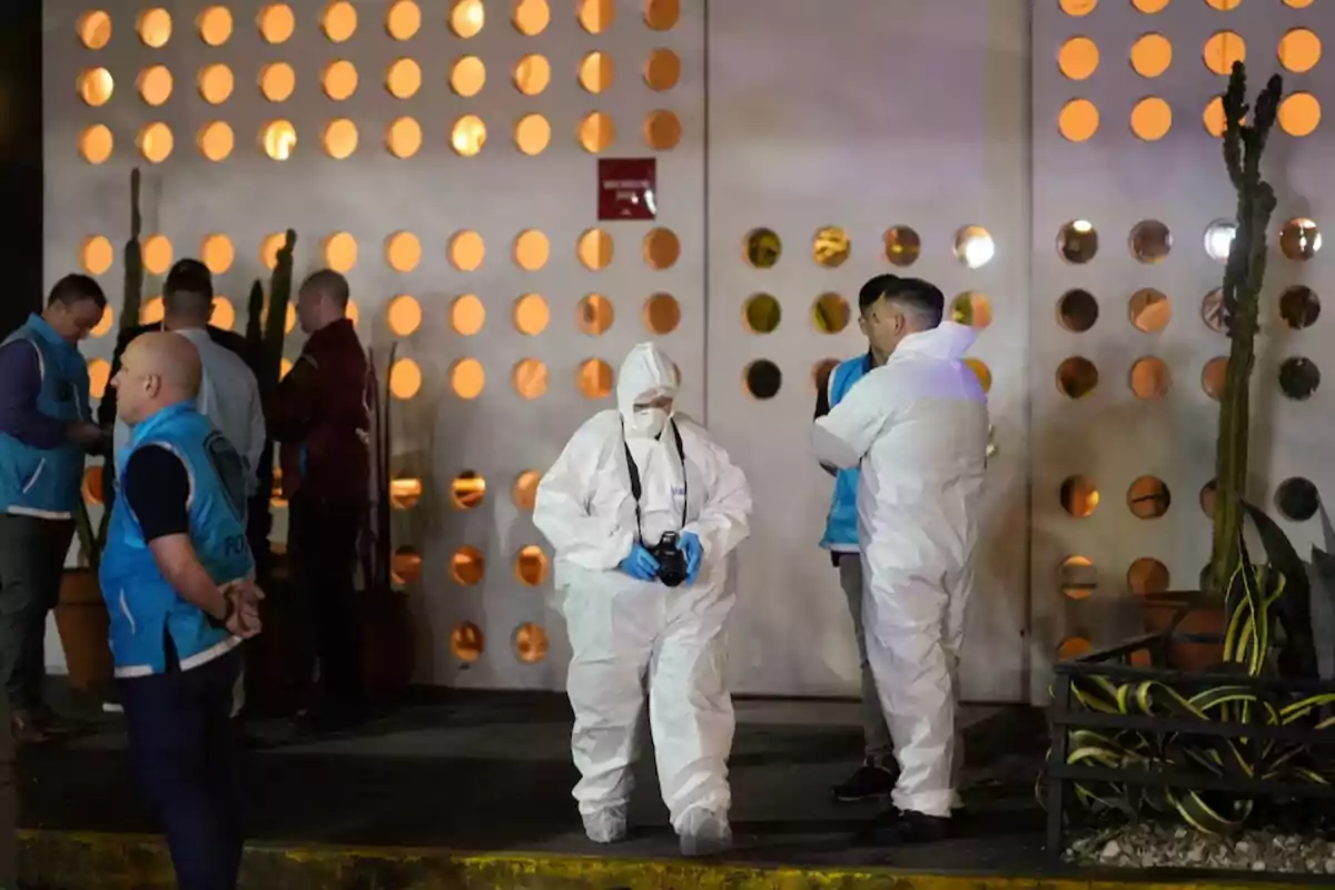 Personas con trajes de protección y chalecos azules se encuentran frente a un edificio con una fachada de círculos iluminados.