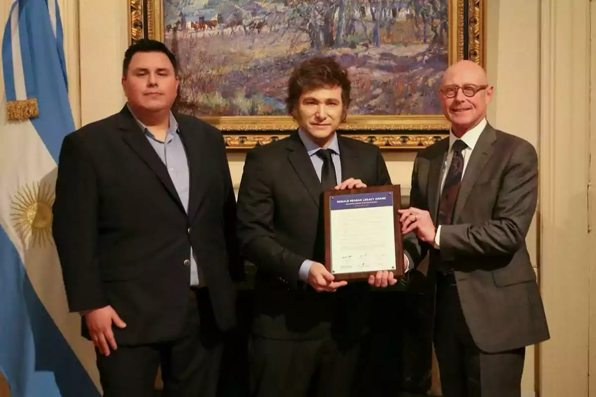 Tres hombres posan juntos en un interior elegante, uno de ellos sostiene un diploma enmarcado, con una bandera argentina y una pintura de fondo.