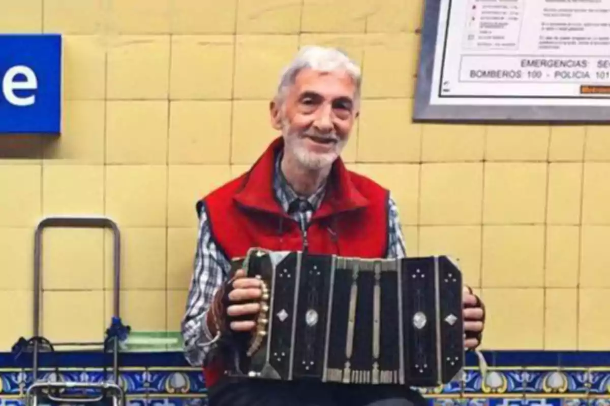 Un hombre mayor con chaleco rojo toca un bandoneón en una estación de metro con azulejos amarillos y un cartel de emergencias en la pared.