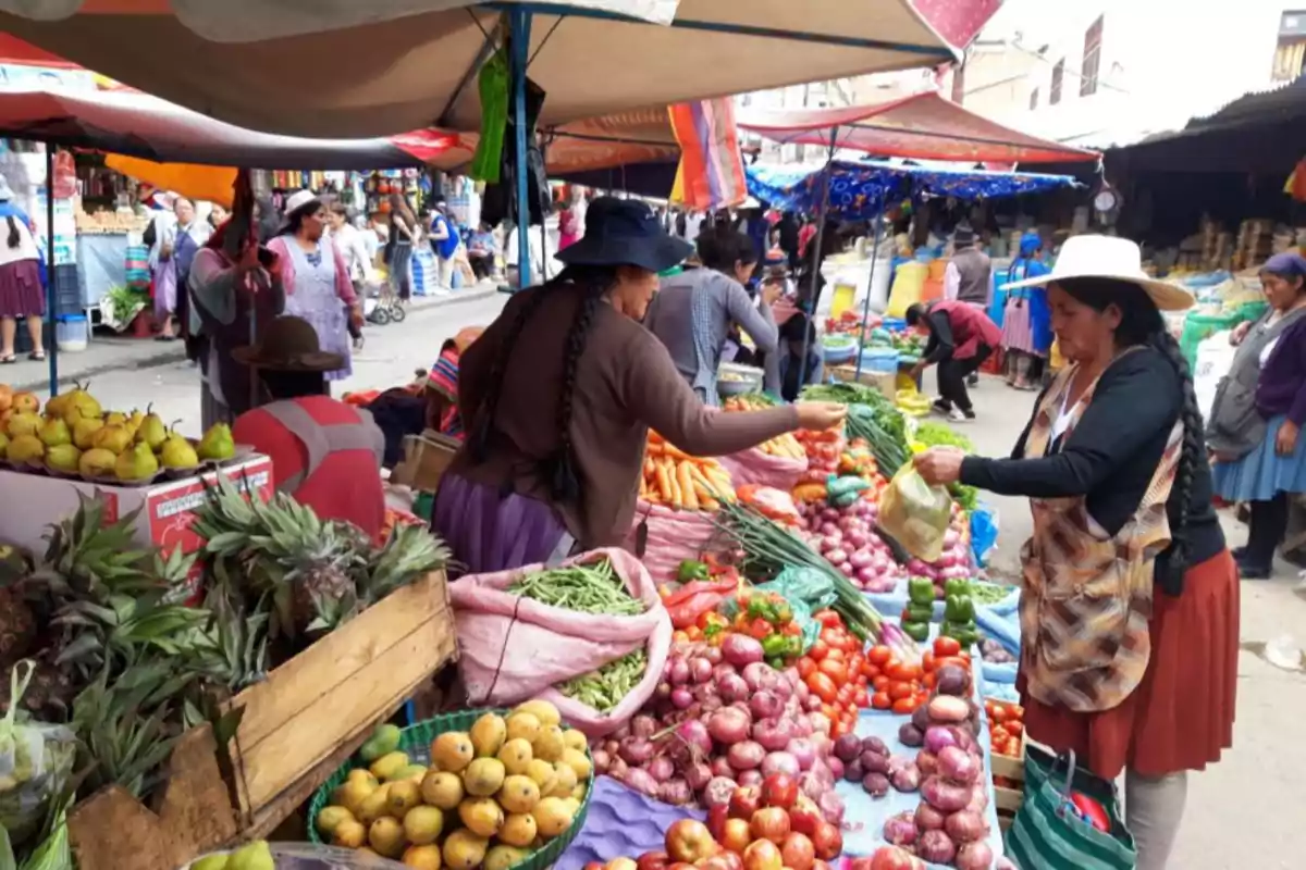 Women buying and selling fruits and vegetables at an outdoor market with stalls full of fresh produce.