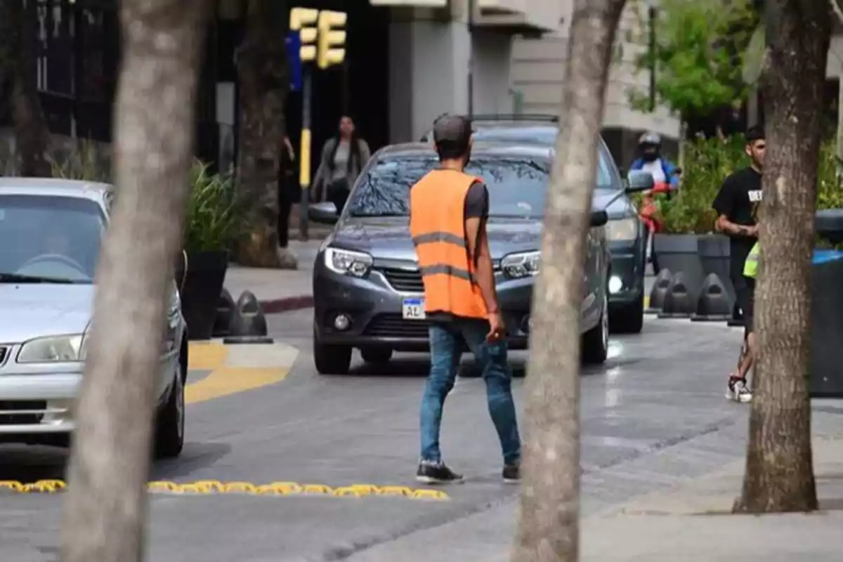Hombre con chaleco naranja caminando entre autos en una calle urbana rodeada de árboles Hombre con chaleco naranja caminando entre autos en una calle urbana rodeada de árboles
