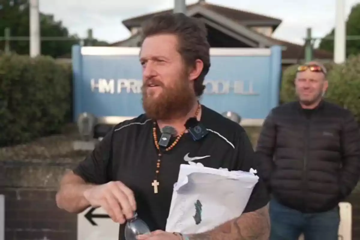 Brown-haired, bearded man holding a bottle and some papers in front of a blue sign with another person in the background Brown-haired, bearded man holding a bottle and some papers in front of a blue sign with another person in the background