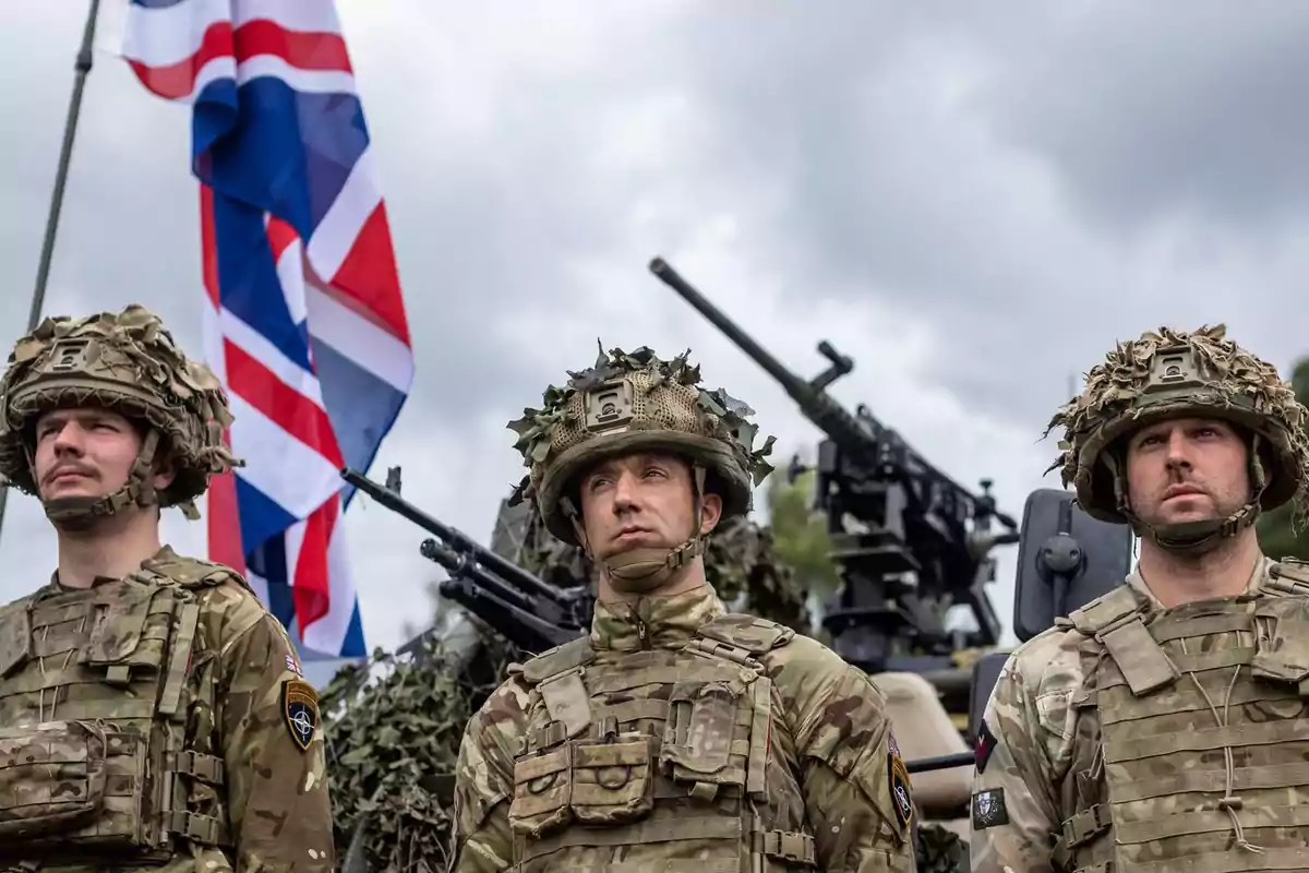 Tres soldados con uniformes de camuflaje y cascos están de pie frente a una bandera británica y equipo militar.