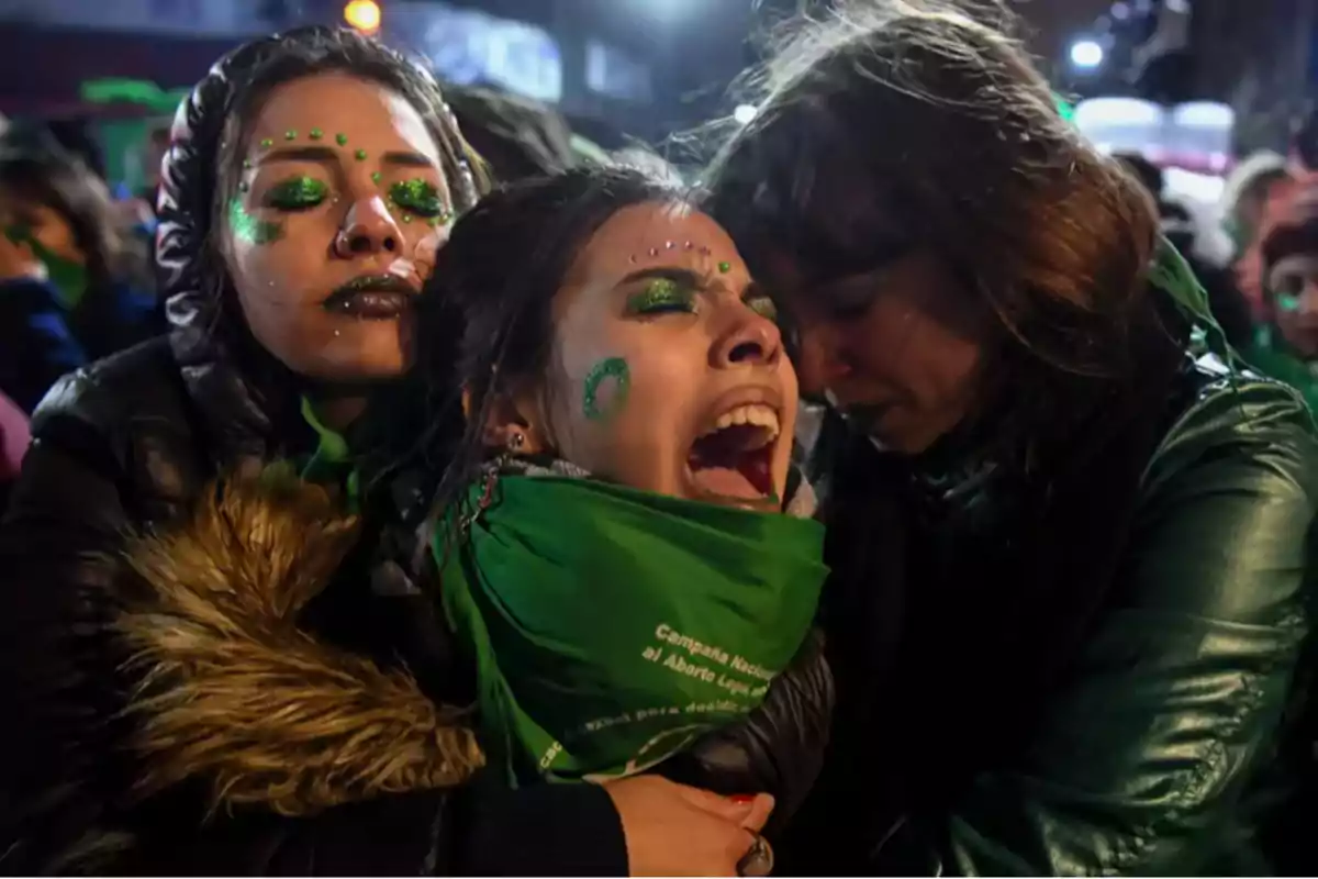 Tres personas con maquillaje verde y pañuelos del mismo color expresan emociones intensas en una manifestación nocturna.